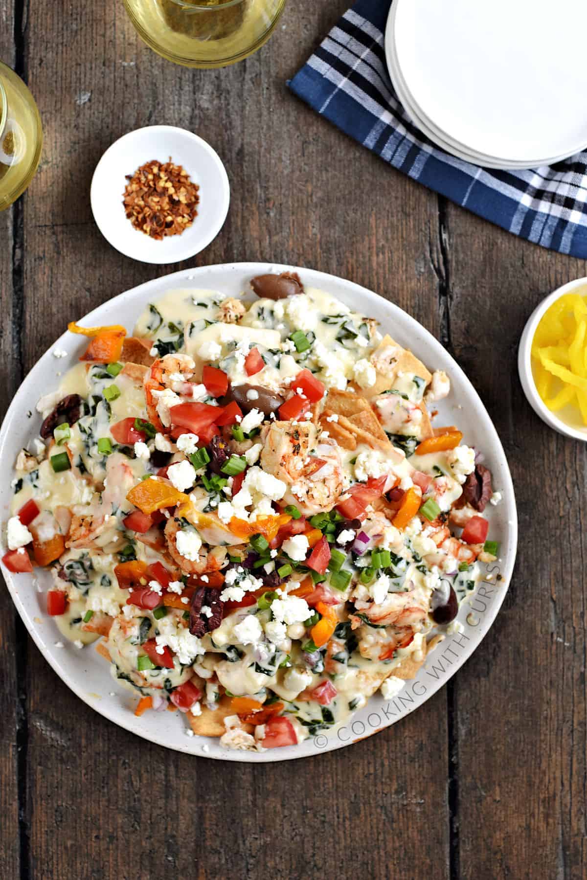 Looking down on a large plate of Greek Seafood Nachos with two glasses of white wine, a bowl of crushed red pepper, and a bowl of banana peppers in the background. 