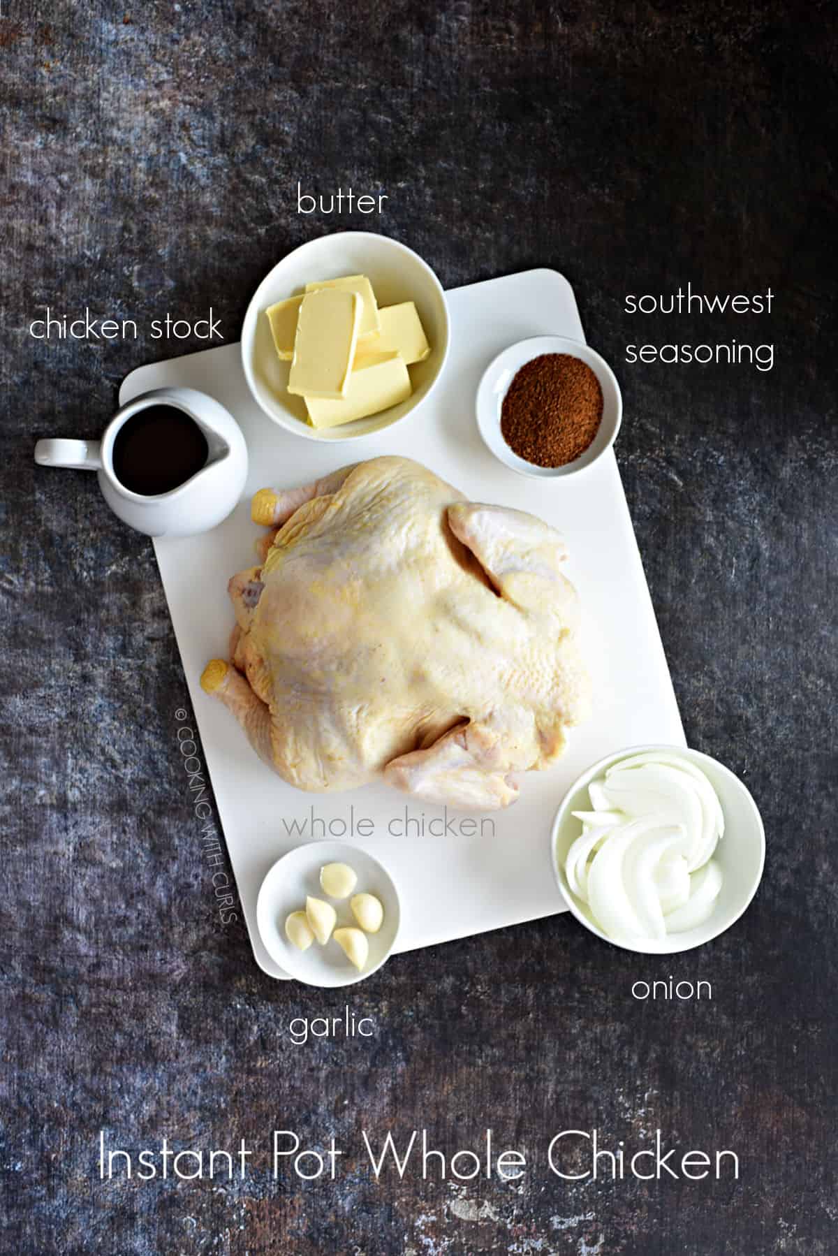 A whole chicken on white cutting board with a pitcher of chicken stock, and bowls of butter, southwest seasoning, sliced onions and garlic cloves. 