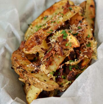 Baked potato wedges topped with Parmesan cheese, bacon, and herbs in a parchment lined basket.