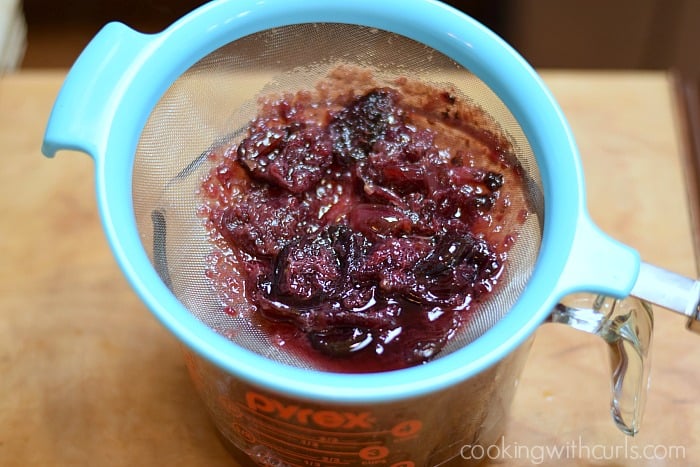 Straining the cherries from the syrup through a blue strainer into a large glass measuring cup.