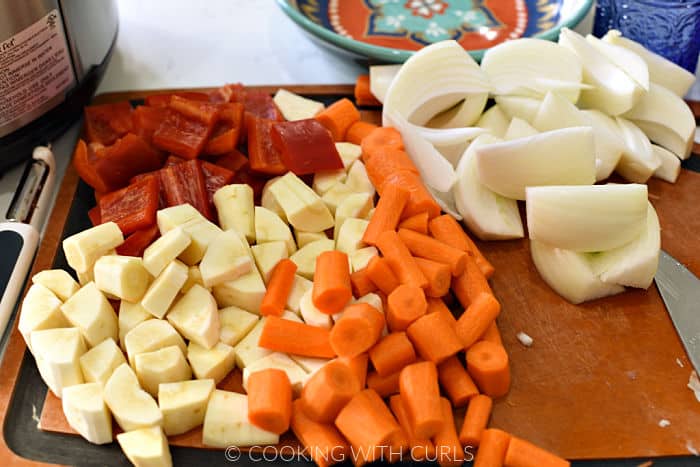 Chopped red bell pepper, parsnip, carrots, and wedged onions on a cutting board. 