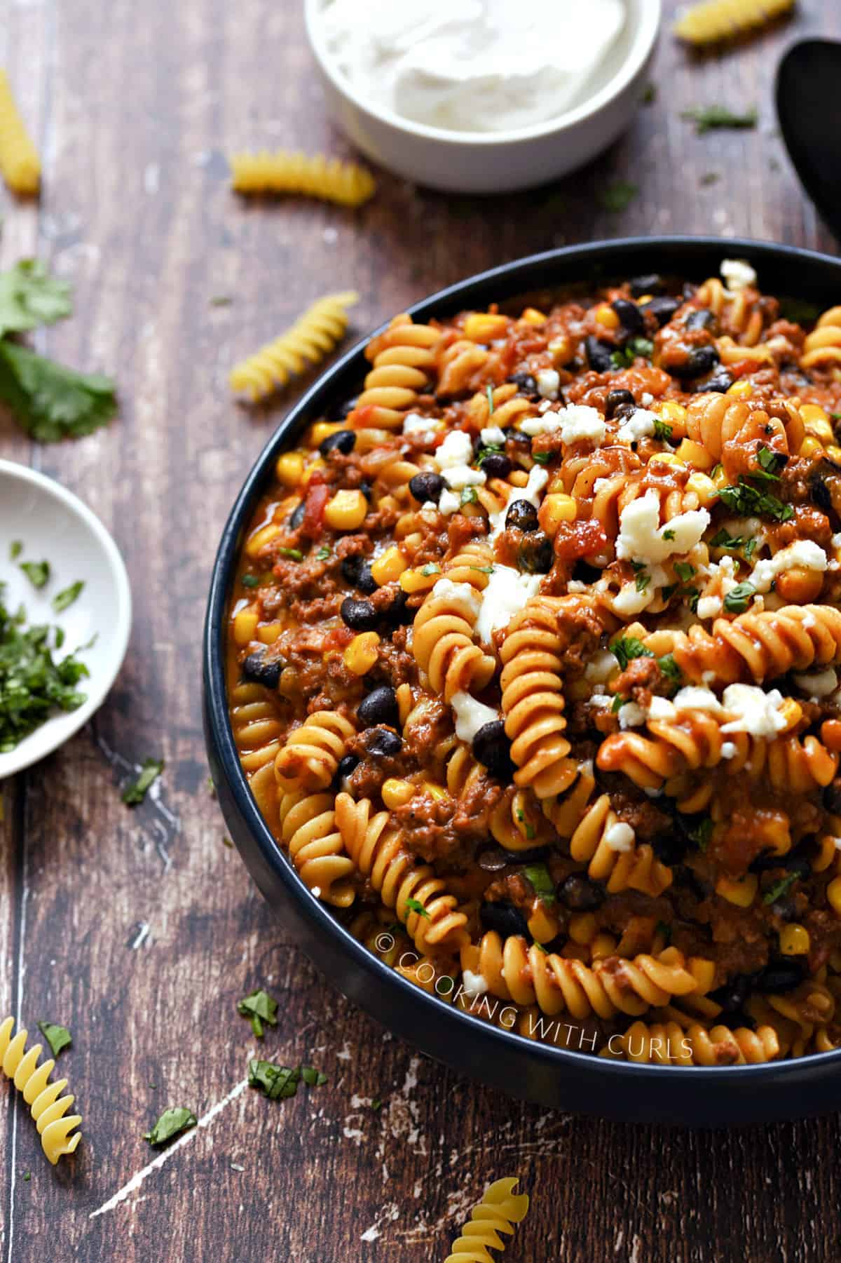 A big blue bowl filled with taco pasta topped with crumbled cheese and surrounded by bowls with sour cream, and chopped cilantro.