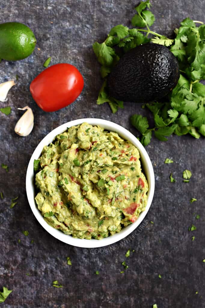 Looking down on a bowl of guacamole with a lime, tomato, garlic cloves and cilantro leaves in the background.