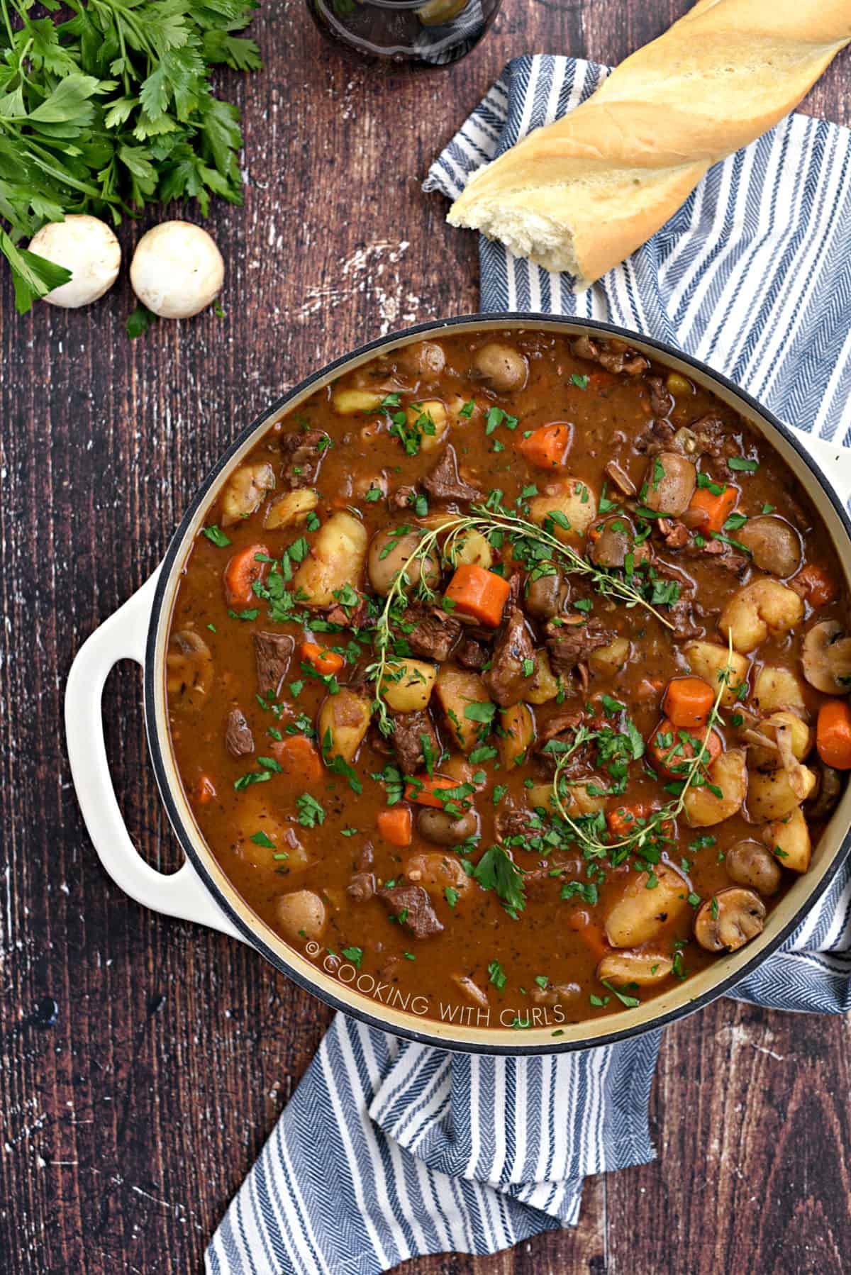 Rich, brown beef stew with chunks of potatoes, carrots, onion, and beef with a baguette, mushrooms, parsley and glass of red wine in the background.