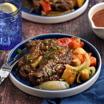 Pot roast with carrots, onions and parsnips on a blue plate with a blue glass and blue napkin in the background.