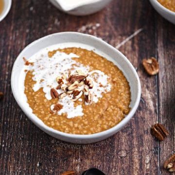 Pumpkin oatmeal in a bowl topped with pecan pieces and cream, with a bowl of cream and second bowl of oatmeal in the background.