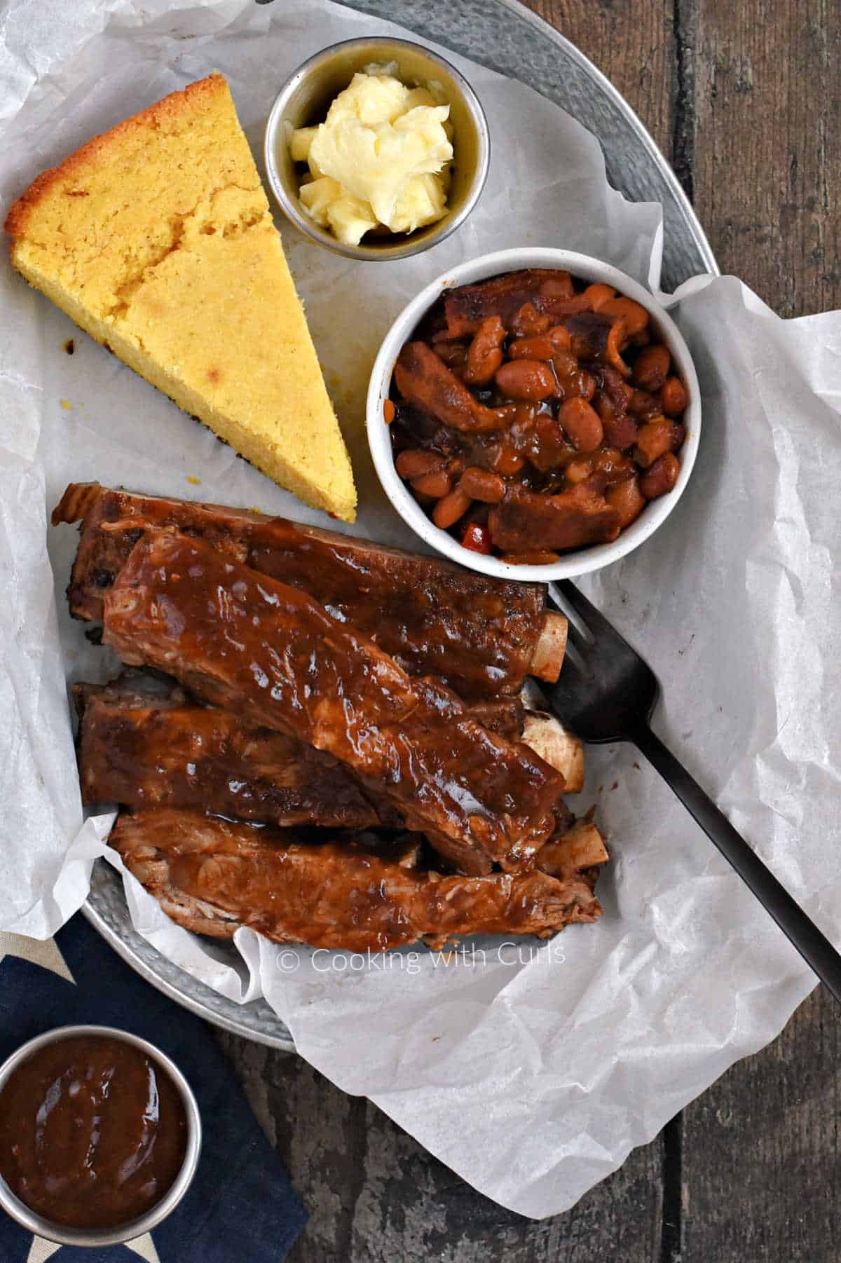 Looking down on a tray with barbecue ribs, a wedge of cornbread, and a small bowl of baked beans with bacon.