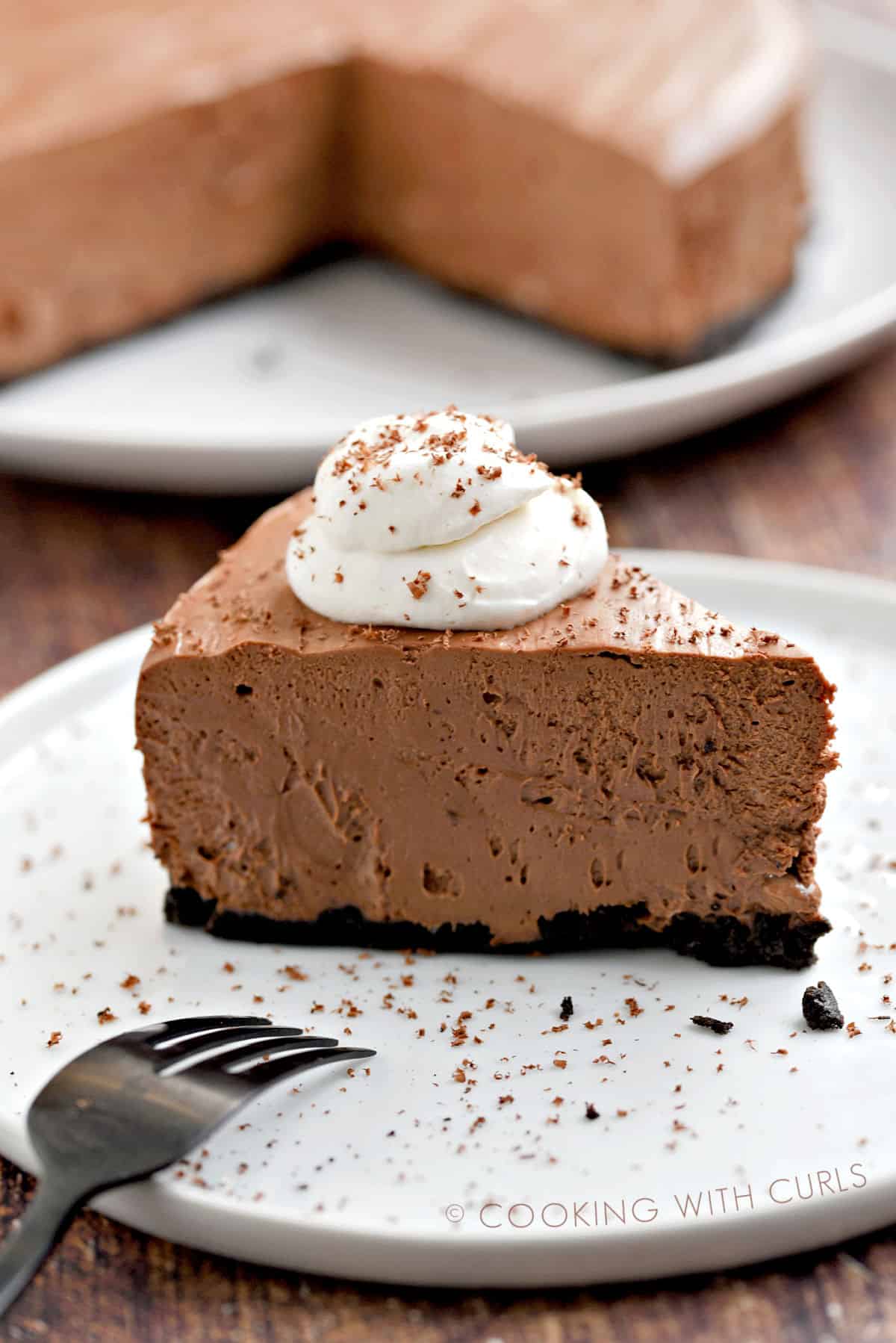 A slice of chocolate cheesecake topped with whipped cream and sprinkled with shaved chocolate sitting on a white plate with the remaining cheesecake on a white plate in the background.