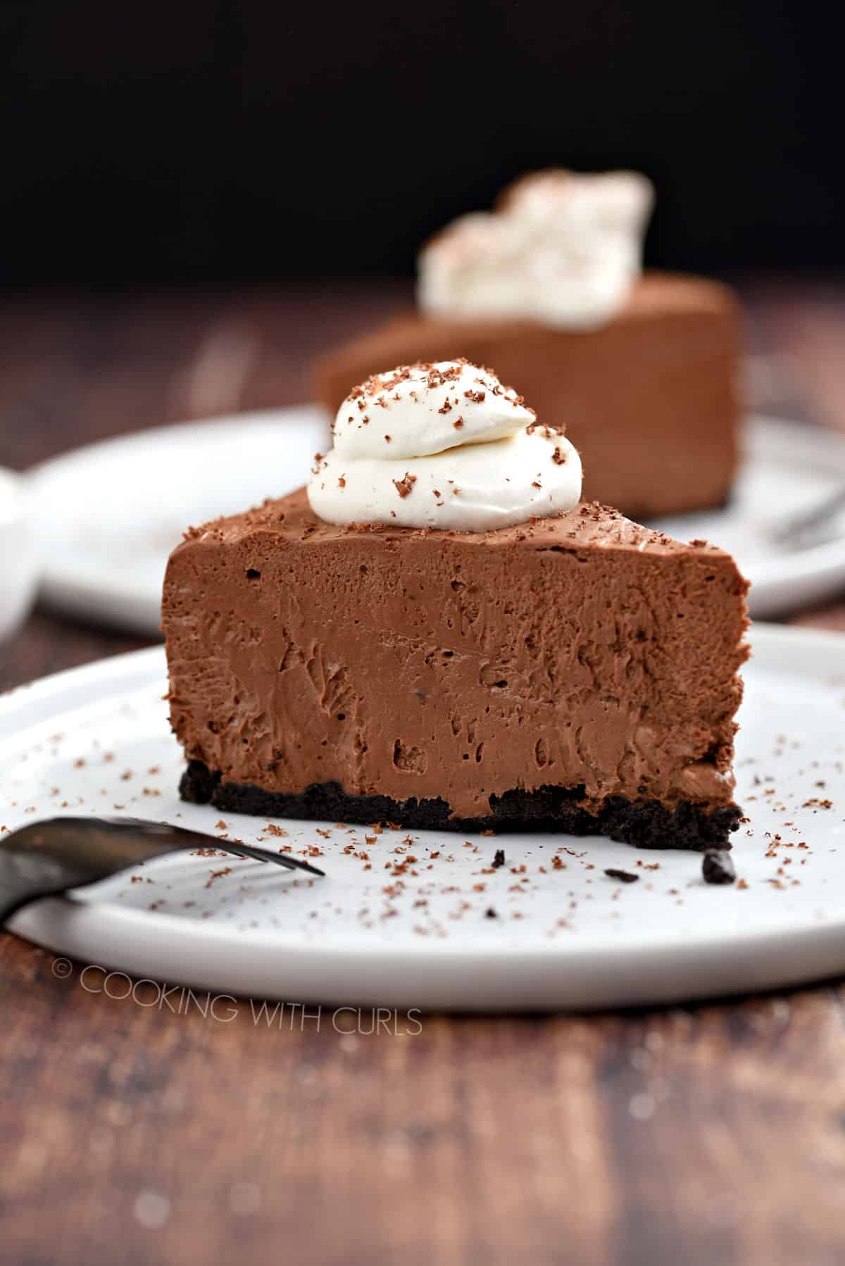 A slice of chocolate cheesecake topped with whipped cream and sprinkled with shaved chocolate sitting on a white plate with a second slice in the background.